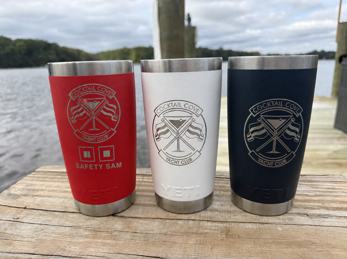 Red Safety Sam, white, and navy 20oz Yeti Ramblers with the Cocktail Cove Yacht Club crest lined up on a dock rail by the water